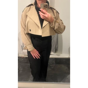 Croppad beige trenchcoat med bälte - Säljer en croppad trenchcoat i beige med bred krage, svarta knappar och snygga bältesdetaljer vid ärmsluten. Jackan har lång ärm och loose passform, perfekt för dig som vill ha en trendig och chill look.