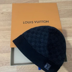 Svart Louis Vuitton mössa - Snygg svart mössa från Louis Vuitton med diskret rutigt mönster och klassisk LV-logga framtill. Mössan har en bredare kant nertill och är tillverkad i mjukt material som håller dig varm, stilren under kalla dagar och äkta 