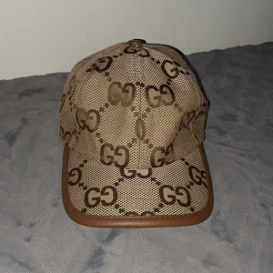 gg keps med mönster beige/brun - Säljer en snygg keps från gg i beige och brunt med det klassiska mönstret över hela kepsen. Kepsen har bruna detaljer och kant i skinnimitation. Perfekt accessoar för att lyfta din outfit. Använd 