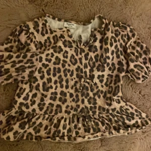 Leopardmönstrad topp 134/140 - Superfin leopardmönstrad klänning från Kappahl i beige och bruna toner. Modellen har volang nertill, korta ärmar och en söt knytning framtill. Perfekt för dig som vill sticka ut med ett djurmönster och gillar trendiga detaljer.