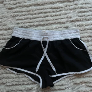 Svarta sportiga shorts med vit kant - Snygga svarta shorts med vit resårmidja och vita detaljer längs kanterna. De har snörning framtill och två fickor med vit piping. Modellen är loose fit och passar perfekt för träning eller chilldagar 🫶🏼 Materialet känns lätt och syntetiskt, vilket gör dem så bekväma och luftiga att bära! Inga tecken på användning. Storlek M men passar både XS och S 💕