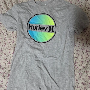 Grå Hurley t-shirt med färgglatt tryck - Snygg grå t-shirt från Hurley i mjuk bomull. Framsidan har ett stort, rundat tryck med blå och limegröna toner samt svart Hurley-logga. Klassisk passform och korta ärmar, perfekt för chill dagar. Enkelt och stilrent plagg med cool färgdetalj.