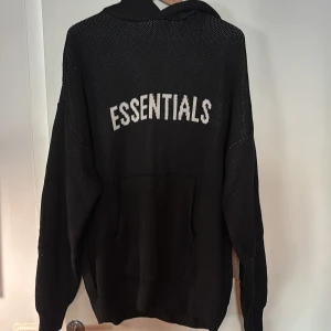 Svart Essentials hoodie stickad XL - Svart stickad hoodie från Essentials Fear of God i storlek XL. Tröjan har huva, stor magficka och vit text 'ESSENTIALS' på ryggen. Tillverkad i 100% bomull och har en loose fit som ger en avslappnad vibe.