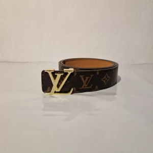 Louis Vuitton brunt skärp med LV-spänne 110 cm - Snyggt brunt Louis Vuitton-bälte med klassiskt monogrammönster och ett stort LV-spänne i guld. Bältet är gjort i exklusivt skinn och har en lyxig känsla som verkligen sticker ut. Perfekt för dig som vill ha en statement-accessoar med ikonisk design.