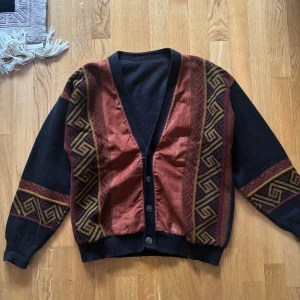 Mönstrad kofta i svart och rost - Unik kofta med svart bas och rostfärgade paneler framtill. Snygga geometriska mönster i gult och vinrött längs ärmar och sidor. Koftan har v-ringning, knappar och är gjord i en mjuk blandning av ull och syntet. Perfekt för dig som gillar retrovibbar och statement-plagg.