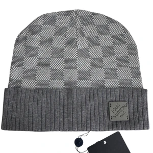 Grå rutig mössa från Louis Vuitton - Snygg grå mössa från Louis Vuitton med klassiskt rutigt mönster och ribbad uppvikt kant. Mössan har en metallbricka med Louis Vuitton Paris framtill och är tillverkad i mjukt material som passar perfekt för kyliga dagar.