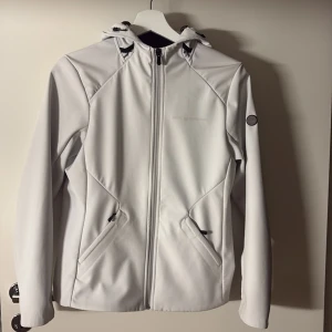 Vit vindjacka från Sail Racing - Snygg vit vindjacka från Sail Racing med huva och hel dragkedja framtill. Jackan har två fickor med dragkedja, svart fleecefoder och diskret logga på ärmen. Perfekt för dig som gillar stilren och sportig look.