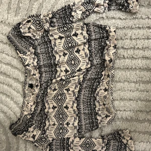 Mönstrad topp från Divided - Topp från Divided med svart och beige etnoinspirerat mönster. Klänningen har långa ärmar och resår i midjan för en snygg passform. Tillverkad i mjuk bomull, perfekt för dig som gillar unika prints och bekväma plagg.