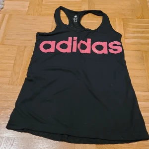 Svart Adidas linnetopp med rosa tryck - Sportig svart linnetopp från Adidas med stort rosa logotryck framtill. Baksidan har diskret prickigt mönster och ett litet rosa Adidas-märke. Modellen är ärmlös med brottarrygg och tillverkad i lätt polyester med Climalite-teknologi.