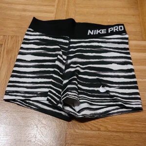 Nike Pro randiga träningsshorts S - Snygga Nike Pro shorts i svart och vitt med randigt mönster. De har en bred elastisk midja med Nike Pro-logga och är tillverkade i ett stretchigt syntetmaterial som sitter tight. Perfekta för gymmet eller sportaktiviteter. Kort modell som ger en sportig vibe.
