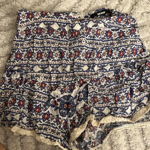 Mönstrade shorts från Bik Bok - Säljer ett par vita shorts från Bik Bok med blått och rött etnoinspirerat mönster. De är högmidjade och har volangdetaljer samt spetskant längst ner. Materialet känns lätt och luftigt, perfekt för varma dagar. Supersnygga till en enkel topp eller linne!