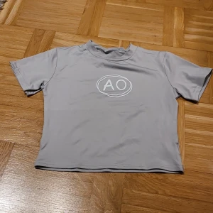 Grå croppad topp med AO-tryck - Snygg ljusgrå croppad topp från AO med korta ärmar och rund hals. Framsidan har ett vitt AO-tryck i en oval. Tillverkad i mjuk och stretchig polyester med elastan för skön passform. Perfekt för dig som gillar stilrena och sportiga plagg.