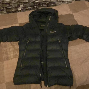 Svart dunjacka från Marmot med huva - Säljer en svart dunjacka från Marmot i storlek S. Jackan har huva, två snedställda dragkedjefickor och en hög krage. Klassisk pufferstil med breda kanaler och diskret logga på bröstet. Perfekt för kalla dagar och riktigt snygg streetstil.