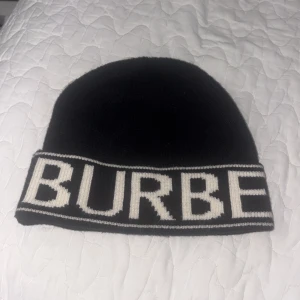 Svart Burberry mössa med logga - Snygg svart mössa från Burberry med bred vit logotyp runt hela kanten. Klassisk och stilren design som passar perfekt till kalla dagar. Mössan är stickad och har en tight passform som sitter skönt på huvudet.
