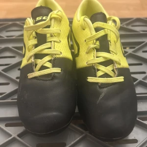 Svarta och gula fotbollsskor med snörning - Säljer ett par fotbollsskor från Nike i svart och gul färg. Skorna har snörning och en rund tå, med slitstarka dobbar under sulan för bra grepp på planen. Ovandelen är tillverkad i syntetmaterial med perforerade detaljer för ventilation. Perfekt för dig som älskar fotboll.