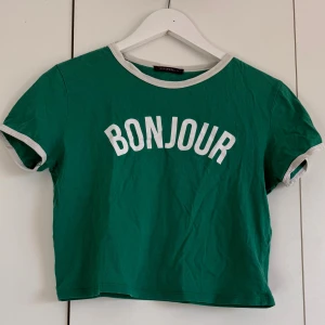 Grön croppad t-shirt med BONJOUR - Säljer en grön croppad t-shirt från Television med vit text 'BONJOUR' framtill. T-shirten har vita kontrastkanter vid halsringning och ärmslut samt korta ärmar. Perfekt för dig som gillar franska vibes och chill stil.