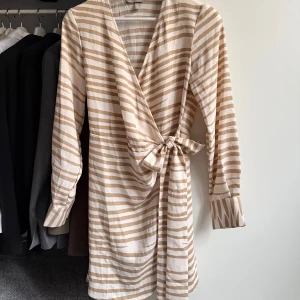 Randig omlottklänning med knyt - 🤍Omlottklänning från H&M i beige och vitt med ränder. Klänningen har långa ärmar, v-ringning och ett knyt i midjan 🤍