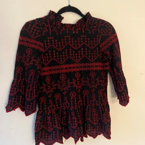 Svart blus med röd brodyr - Unik svart blus med röd broderad design och volangdetaljer. Blusen har trekvartsärmar, hög krage med rysch och ett vackert mönster över hela plagget. Perfekt för dig som vill sticka ut med en cool och bohemisk vibe.