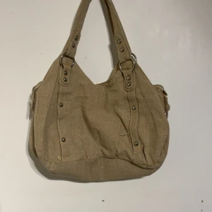 Beige axelväska från KappAhl  - Stilren beige axelväska från KappAhl i linnematerial med snygga metalldetaljer och ringar på handtagen. Väskan har ett glansigt innerfoder i satin och praktisk dragkedja. Perfekt för dig som vill ha en enkel men trendig väska med mycket plats.