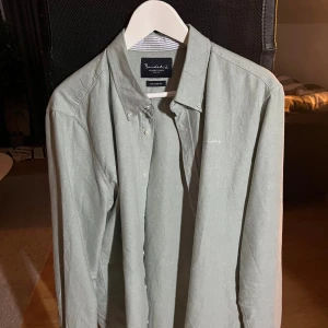 Ljusgrå skjorta från Bondelid - Snygg ljusgrå skjorta från Bondelid med klassisk krage och diskret broderad logga på bröstet. Skjortan har knappar framtill och en stilren look, perfekt för dig som gillar enkel och clean stil. Materialet är mjukt bomullstyg och insidan av kragen har tunna blå ränder.