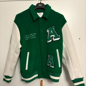 Grön varsityjacka Axel Arigato - Snygg grön och vit varsityjacka från Axel Arigato med stora broderade bokstäver och detaljer på både fram- och baksida. Jackan har vita ärmar, grönt kroppsparti, ribbade muddar och knappar framtill. Perfekt för dig som vill sticka ut med en sportig och trendig look.