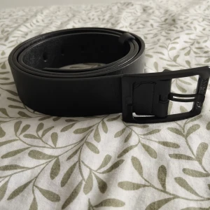 Svart skärp - Stilrent svart bälte från ZOORLULANN med matt svart fyrkantigt spänne där loggan är ingraverad. Bältet är i slät skinnimitation och har en klassisk rak form. Perfekt för att ge din outfit en clean och modern touch.