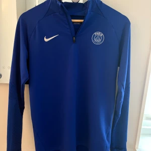 Blå Nike PSG half zip träningströja - Snygg blå långärmad träningströja från Nike med Paris Saint-Germain-logga på bröstet. Tröjan har half zip framtill, vita Nike-swoosh och diskret mönster på ärmarna. Slim fit och tillverkad i polyester för skön känsla under fotbollsträningen.
