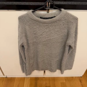 Grå fluffig stickad tröja - Mysig grå stickad tröja med fluffig yta och rund hals. Perfekt för dig som gillar en enkel men ändå trendig look. Långärmad modell med relaxed passform som är skön att bära under kyliga dagar.