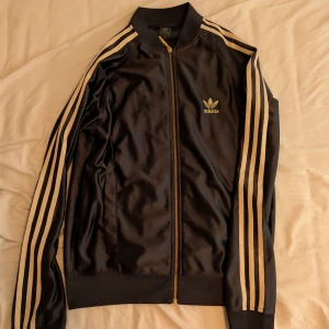 Svart Adidas bomberjacka med guld - Snygg svart teöja från Adidas med klassiska guldiga ränder längs ärmarna och guldfärgad dragkedja. Jackan har Adidas logga broderad på bröstet och ribbade muddar vid ärmslut och nederkant. Perfekt för en sportig och trendig look.