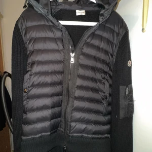 Moncler cardigan svart pufferjacka med stickade ärmar och huva - Snygg svart cardigan från Moncler med stickade ärmar och huva. Jackan har quiltad front, dragkedja och en praktisk ficka med dragkedja på ärmen. Perfekt för dig som vill ha en sportig och stilren look med exklusiv premiumkänsla.