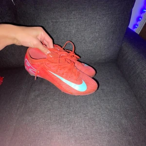 Ett par röda Nike Vapor 16 elite 42 - Säljer ett par snygga Nike fotbollsskor i röd färg med turkos swoosh på sidan. Skorna har snörning och en platt sula med dobbar för bra grepp på planen. Ovandelen är i syntetmaterial och designen är sportig med coola detaljer på sulan. Perfekt för dig som vill sticka ut på matchen. De kommer med original lådan