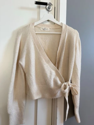 Beige omlottkofta från H&M - Mysig beige omlottkofta från H&M med knytband i midjan och ribbade muddar.