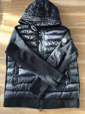 Moncler Cardigan - Snygg svart dunjacka från Moncler med stickade ärmar och huva. Jackan har dragkedja framtill, två sidofickor och en Moncler-logga på ärmen. Perfekt för en stilren look. Storlek L men passar M