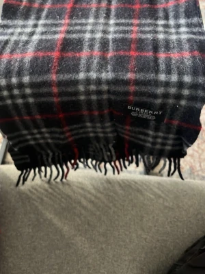 Rutig halsduk från Burberry i ull - Snygg halsduk från Burberry i 100% lammull med klassiskt rutmönster i svart, grått, vitt och rött. Halsduken har fransar i ändarna och är tillverkad i England. Perfekt accessoar för att lyfta din outfit.