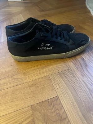 Svarta Saint Laurent sneakers - Säljer ett par svarta Saint Laurent sneakers med vit logga på sidan. Skorna har lågt skaft, svarta skosnören och en beige sula. Materialet är canvas och mocka vilket ger en stilren och klassisk look. Perfekta för dig som gillar exklusiva märken och enkel design.
