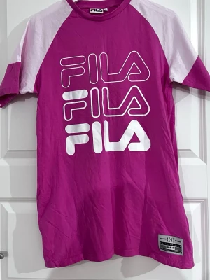 Rosa och vit FILA t-shirtklänning - Snygg rosa t-shirtklänning från FILA med vita kontrastärmar och stort FILA-tryck framtill. T-shirten har klassisk passform, rund hals och är tillverkad i mjuk bomull. Perfekt för dig som gillar sportig och trendig stil. 2 fickor. 