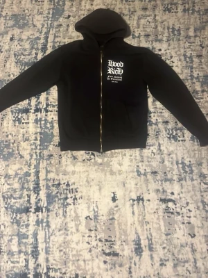 Svart zip hoodie från Hoodrich - Säljer en svart hoodie med dragkedja från Hoodrich. Tryck i vitt på bröstet och stort grafiskt tryck på ryggen. Klassisk huva och känguruficka framtill. Perfekt för dig som gillar streetwear och vill sticka ut med coola detaljer.