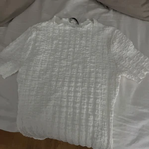 Zara topp - Säljer en vit smockad topp med kort ärm och rund hals. Toppen har en tight passform och är gjord i ett stretchigt, strukturerat material som ger en cool textur. Perfekt att styla med kjol eller jeans för en fräsch look.
