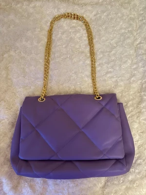Lila quiltad handväska med guldkedja - Säljer en snygg lila handväska med quiltat mönster och en lång guldfärgad kedjerem. Går att ha i två olika längder (se bilder). Köpt begagnad men i fint skick, skulle inte tro att den är använd så mycket. Bara att skriva om du undrar nåt eller vill ha mått💕