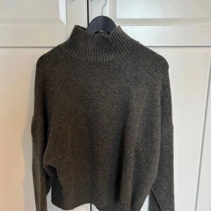 Mörkgrå stickad polotröja - Mysig mörkgrå stickad polotröja med bred ribbad krage och muddar. Tröjan har en avslappnad passform och är perfekt för kyliga dagar. Materialet ser ut att vara ullblandning och modellen är något boxig med långa ärmar.