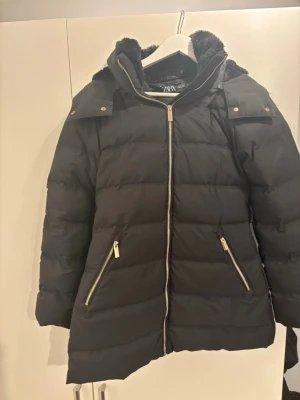 Svart dunjacka med fuskpäls från Zara - Svart dunjacka från Zara med avtagbar huva och fuskpäls. Jackan har två snedställda fickor med dragkedja i guld och quiltad design. Perfekt för vintern med sin tjocka vaddering och snygga detaljer.