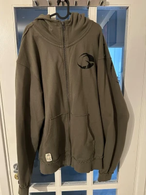 Olivgrön zip-hoodie från Gasp - Säljer en olivgrön hoodie med dragkedja från Gasp. Hoodien har stor huva, känguruficka och tryck med Gasp-logga på bröstet och stort tryck över ryggen. Perfekt för dig som gillar streetwear och sportig stil.