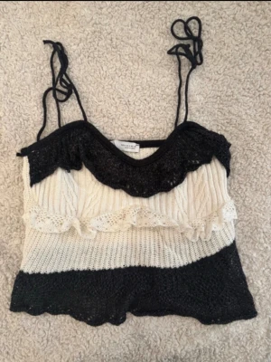 Svartvit stickad linnetopp med volang - Snygg stickad linnetopp från Zara i svart och vitt med volangdetaljer och knytbara axelband. Toppen är perfekt för en trendig look. Materialet är mjukt och luftigt, vilket gör den skön att bära under varma dagar.