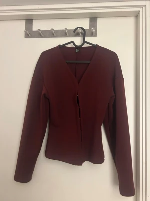 Vinröd cardigan med v-ringning - Vinröd cardigan med v-ringning och knäppning framtill. Långärmad och figurnära modell, perfekt att styla över en topp eller klänning. Enkel och stilren design som passar till många olika looks.