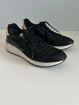 Tamaris sneakers i äkta läder - Snygga svarta sneakers från Tamaris i äkta skinn /läder. Storlek 40. Fina detaljer i guld och i högblank läderprägling. Bekväm vit sula och praktisk guldig dragkedja bredvid den klassiska snörningen så man kan välja. Riktiga kvalitetsskor. Mycket bra skick. Nypris 999:-    Hämtas i Stockholm eller skickas mot porto. 