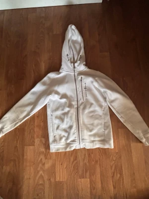 Vit hoodie från Sail Racing - Snygg vit hoodie från Sail Racing med dragkedja framtill och broderad logga på bröstet. Hoodien har två fickor och en skön huva. Perfekt för dig som gillar stilren och sportig look. Materialet känns mjukt och bekvämt mot huden.