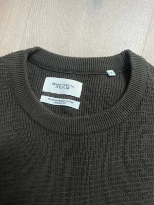 Olivgrön stickad tröja Marc O'Polo - Olivgrön stickad tröja från Marc O'Polo Denim i premium ekologisk bomull. Tröjan har rund halsringning, lång ärm och ett snyggt strukturerat mönster. Storlek M