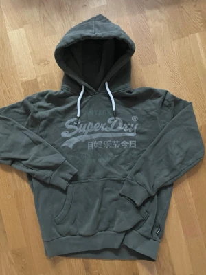 Grön hoodie från Superdry - Snygg grön hoodie från Superdry med stor logga och japansk text på bröstet. Klassisk känguruficka framtill, vita dragsnören i huvan och ribbade muddar vid ärmar och nederkant. Perfekt för en avslappnad och cool stil.