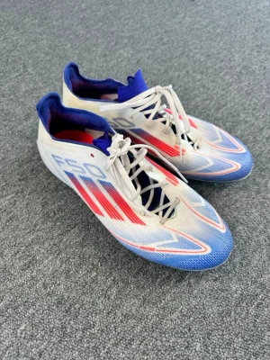 Adidas F50 fotbollsskor blå/röd/vit - Säljer ett par Adidas F50 fotbollsskor i vitt med blå och röda detaljer. Skorna har snörning, låg profil och mönstrad sula med fasta dobbar för gräsplan. Snygg design med klassiska tre ränder och F50-tryck på sidan. Perfekta för dig som vill sticka ut på planen.