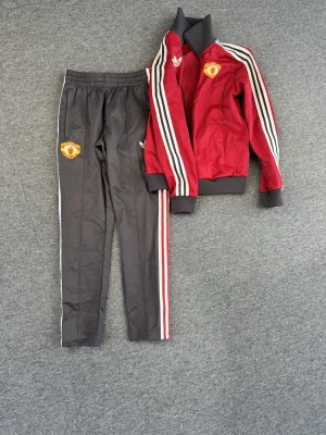 Manchester United Adidas tracksuit  - Snyggt träningsset från Adidas och Manchester United. Jackan är röd med svarta detaljer, vita och röda ränder på ärmarna samt klubbmärke på bröstet. Byxorna är svarta med klubbmärke och matchande ränder längs benen. 